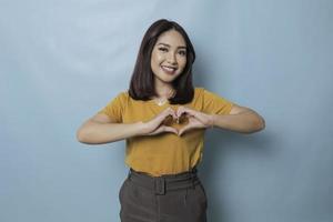 Attractive young Asian woman feels happy and romantic shapes heart gesture expresses tender feelings wears casual yellow t-shirt against blue background. People affection and care concept photo