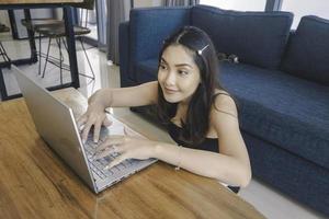 la joven asiática pasa su tiempo en casa sentada en la sala de estar sonriendo y trabajando en su computadora portátil. foto