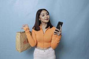 retrato asiático feliz hermosa mujer joven de pie emocionado sosteniendo una bolsa de compras en línea y un teléfono inteligente en la mano, foto de estudio aislada en el fondo azul