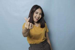 Attractive young Asian woman feels happy and romantic shapes heart gesture expresses tender feelings wears casual yellow t-shirt against blue background. People affection and care concept photo