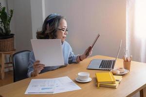 Portrait of an elderly woman working inside the house using laptops and tablets to work and check important documents for customers. photo