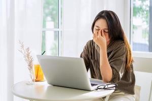 Tired and stressed young Asian woman feel pain eyestrain and rubbing her eyes and take off her glasses while sitting at her working place photo