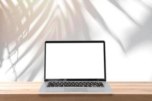 Empty screen laptop on a wooden table with shadow of palm leave on white cement wall background and copy space photo