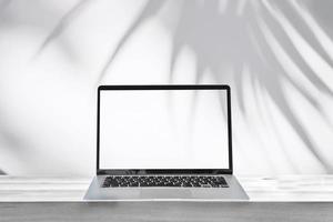 Empty screen laptop on a wooden table with shadow of palm leave on white cement wall background and copy space photo