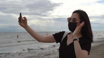 jovem mulher asiática usando máscara médica e videochamada ou facetime por smartphone na praia após coronavírus, novo estilo de vida normal e conceito de bolha de viagem