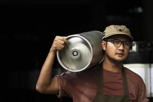 Young man in leather apron holding beer keg at modern brewery, craft brewery worker photo