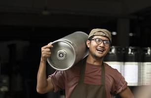 Young man in leather apron holding beer keg at modern brewery, craft brewery worker photo