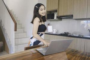 Young Asian woman spends her time at home working remotely in the dining room having an online conference on her laptop while carrying a mug. photo