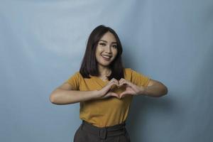 Attractive young Asian woman feels happy and romantic shapes heart gesture expresses tender feelings wears casual yellow t-shirt against blue background. People affection and care concept photo