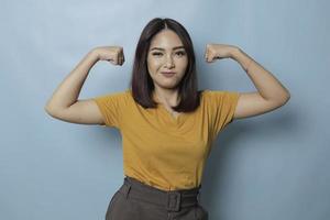 joven hermosa chica con una camiseta amarilla informal de pie sobre un fondo azul aislado que muestra los músculos de los brazos sonriendo orgulloso. concepto de fitness. foto