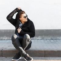 Young stylish guy in sunglasses sitting on the curb on the background of a white wall. photo