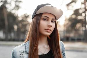 Close-up portrait of a beautiful hipster girl in a black baseball cap and torn blue jeans jacket at sunset photo