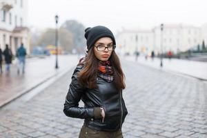 moda look.glamour modelo de mujer morena de estilo de vida en chaqueta de cuero negro y una gorra de punto negro al aire libre en la calle. foto