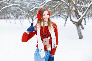 hermosa joven en un suéter rojo con un reno y auriculares de invierno para divertirse en el parque. mostrando pulgares arriba signo de calidad foto