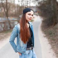 Fashionable young girl in a black cap and jeans jacket standing on the background of the spring park photo