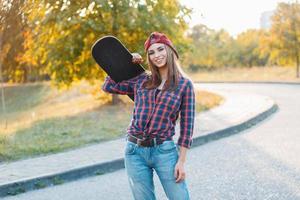 hermosa joven hipster con ropa elegante sosteniendo una patineta y sonriendo al atardecer foto