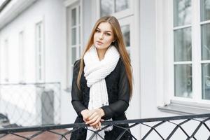 Beautiful girl with a white knitted warm scarf and a black dress stands near home photo