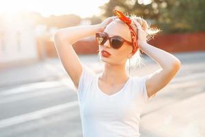 Beautiful hipster woman with red lips and sunglasses standing in the street at sunset photo