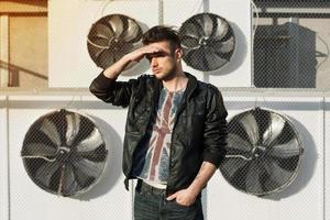 Beautiful guy in a black leather jacket. Stands near air conditioners. Looks into the distance. photo