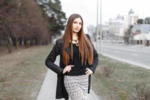 Beautiful young girl with long hair in a stylish black coat posing on the background of the city. photo