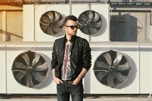 retrato de un hombre hipster con una chaqueta de cuero negra y gafas de sol. se encuentra cerca de acondicionadores de aire. foto
