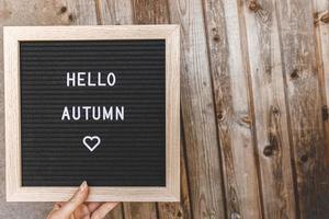 Autumnal Background. Woman hand holding black letter board with text phrase Hello Autumn on wooden palnks backdrop. Thanksgiving banner. Hygge mood, cold weather concept photo