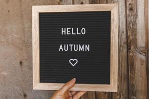 Autumnal Background. Woman hand holding black letter board with text phrase Hello Autumn on wooden palnks backdrop. Thanksgiving banner. Hygge mood, cold weather concept photo