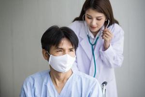 Asian woman doctor use a stethoscope to check the lung rhythm of a male patient who wear face mask while he sit on a wheelchair at hospital. photo