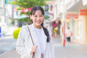 Confident young business Asian working female who wears a white shirt and shoulder bag smiles happily while she is walking to work at office in the city. photo