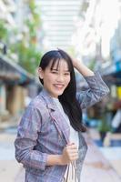 Confident professional young Asian business woman who wears a brown striped blazer and shoulder bag smiles happily and looks at the camera as she commute to work through the old town. photo