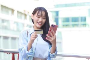 Professional Asian woman in a blue shirt smiles happily successful outdoors in the city while holds smartphone and credit card among the building as a background. photo