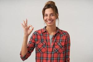 Portrait of cheerful pretty woman in checkered shirt with casual hairstyle showing ok gesture, smiling widely to camera, posing over white background photo