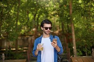 Surprised attractive young bearded man in sunglasses standing over green park with mobile phone photo