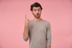 Indoor photo of dark haired bearded male with astonished face posing over pink background, wearing grey sweater, looking at camera and raising eyebrows, pointing upwards with index finger