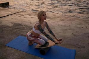 Sporty young pretty woman with good body shape sitting on sports mat and making exercise with balance board, posing over sea view during sunrise, wearing sporty clothes photo