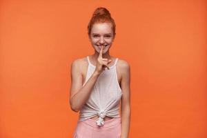 Positive beautiful young lady with foxy hair combed in bun wearing white top and pink skirt, raising forefinger to her mouth in hush gesture, smiling to camera over orange background photo