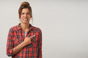 Indoor shot of young female showing with forefinger aside, wearing checkered shirt and natural make-up, pouting and frowning, showing disgust on her face photo