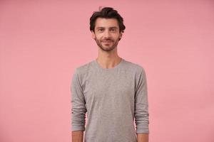 Indoor shot of bearded male with trendy haircut standing over pink background, looking at camera with light smile, wearing grey sweater, showing positive attitude photo