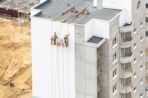 los trabajadores en una grúa pintan la pared de un edificio residencial de gran altura desde la vista de un pájaro con un bosque en el fondo foto