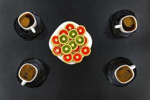 four cups of morning coffee and a plate of colored cookies on a black wooden table. Top view photo