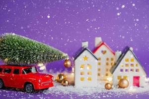 Christmas decor - red retro car on snow carries past houses with fairy lights in bokeh Christmas tree with gift boxes on roof. Toy on violet background. New Year greeting card. Cozy home photo