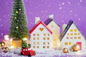 decoración de navidad - coche retro rojo en la nieve pasa por casas con luces de hadas en bokeh árbol de navidad con cajas de regalo en el techo. juguete sobre fondo violeta. tarjeta de felicitación de año nuevo. hogar acogedor foto