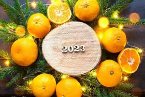 New year's holiday background on a round cut of a tree surrounded by tangerines, live fir branches and golden lights garlands, with wooden numbers date 2023. Citrus aroma, Christmas. Space for text. photo