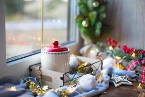 Christmas decor by the window on a cozy wooden windowsill with a mug with a drink and a cookie jar. Fairy lights, Christmas tree branch, breakfast tray still life photo