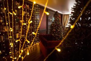 interior navideño de la oscura sala de estar de la casa. árbol de navidad, luces de guirnaldas en las paredes, mueble biblioteca, libros en los estantes. interiores de lujo. año nuevo, bolas rojas y doradas. foto