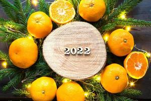 New year's holiday background on a round cut of a tree surrounded by tangerines, live fir branches and golden lights garlands, with wooden numbers date 2022. Citrus aroma, Christmas. Space for text. photo