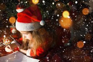 Little girl in a Santa hat and red dress under Christmas tree is dreaming, waiting for the holiday, lying on a plaid blanket. A letter on piece of paper, gifts. New year, Christmas. defocus lights photo