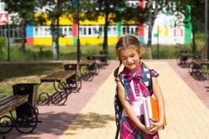 chica con mochila y una pila de libros cerca de la escuela. regreso a la escuela, el niño está cansado, pesados libros de texto. educación, clases de escuela primaria, el comienzo del año escolar, 1 de septiembre foto