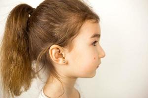 Ear piercing in a child - a girl shows an earring in her ear made of a medical alloy. White background, portrait of a girl with a mole on his cheek in profile. photo