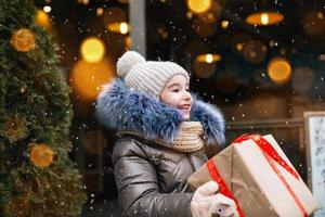 retrato de niña alegre con una caja de regalo para navidad en una calle de la ciudad en invierno con nieve en un mercado festivo con decoraciones y luces. ropa de abrigo, gorro de punto, bufanda y piel. copie el espacio